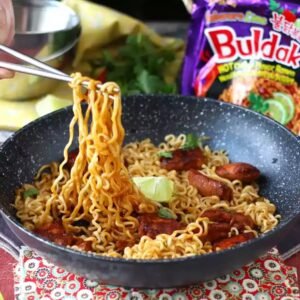 Cooked Samyang Buldak Habanero Lime noodles being lifted with metal chopsticks from a black frying pan with lime wedges and cilantro.