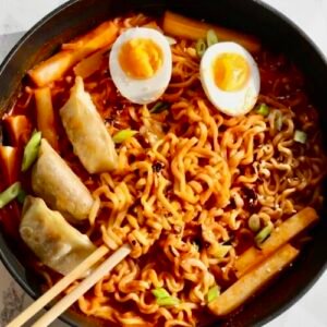 Top-down view of a bowl of spicy Samyang Buldak noodles topped with two halved boiled eggs, pan-fried dumplings, and green onions.