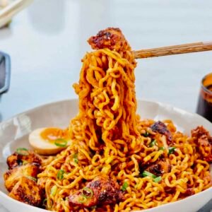 Close-up of spicy Samyang Buldak noodles being lifted with wooden chopsticks, topped with sliced green onions, sesame seeds, and a soft-boiled ramen egg.